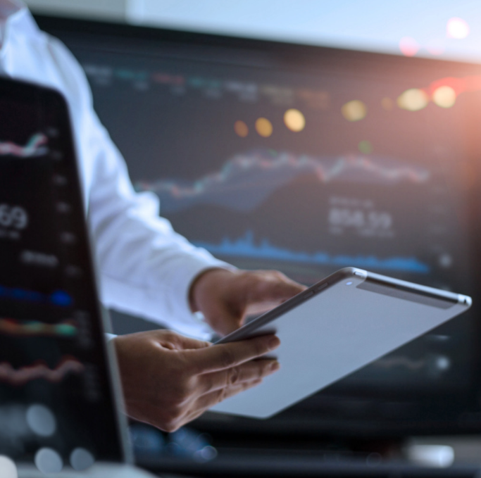 Foto de uma pesoa segurando um tablet, com um notebook mostrando um gráfico na frente da pessoa e uma tela de televisão atras mostrando infromações de gráficos.
